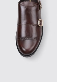 Zapato de cuero marrón con puntera redondeada, cierre con una hebilla, perforaciones decorativas y suela negra. Textura suave.