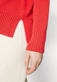 Rode gebreide trui met geribde manchetten en een zijsplit, gecombineerd met een wit kledingstuk. De textuur lijkt zacht en glad.