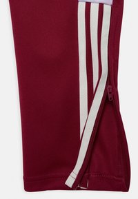 Maroon athletic pants with white accent stripes and a zippered side opening. Made of textured fabric, featuring a snug fit and clean stitching.
