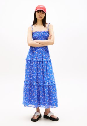 Kvinde står med armene krydsede iført en blå blomsterprint maxi-kjole, rød kasket og hvide platformssandaler på en hvid baggrund.