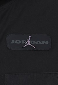 Patch di tessuto nero con la scritta "JORDAN" in lettere in rilievo, accompagnata da un logo silhouette viola sopra, su uno sfondo testurizzato.