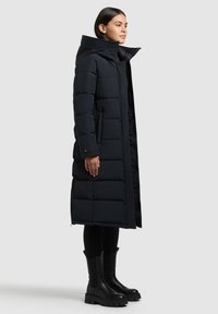 Abrigo de plumas negro largo con un alto cuello, diseño acolchado y bolsillos laterales, combinado con botas negras hasta la rodilla y leggins negros ajustados.