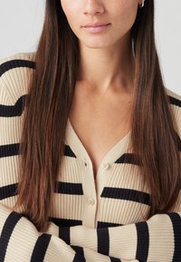 Beżowy prążkowany kardigan z czarnymi poziomymi paskami, dwoma guzikami przy dekolcie w serek, długimi rękawami i miękką fakturą.