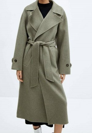 Woman wearing a long, belted olive-green wool coat with wide lapels and buttoned cuffs over a black outfit.