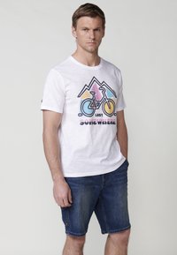 White cotton t-shirt featuring a colorful bicycle graphic and mountain design, paired with blue denim shorts. Simple, casual look.