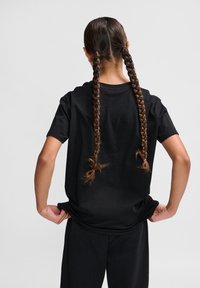 Tricou negru din bumbac cu mâneci scurte, având o croială lejeră și un guler rotund. Părul este coafat în două împletituri lungi.