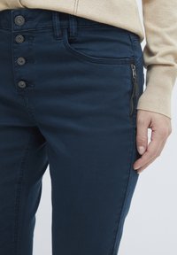 Marineblauwe broek met een ritssluiting zak, sluiting met vijf knopen en zichtbare stikseldetails. Het materiaal lijkt glad met een lichte glans.