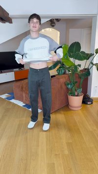 Γκρι και λευκή κοντή μπλούζα με το κείμενο "ΑΣ ΜΕΤΑΤΡΑΠΟΥΜΕ", συνδυασμένη με μαύρο παντελόνι ρίγας και λευκά παπούτσια, σε εσωτερικό περιβάλλον.