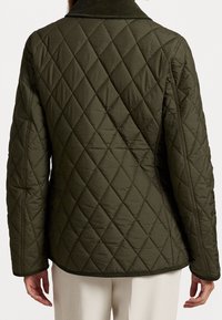 Persoon in een olijfgroene gewatteerde jas met ruitpatroon, gezien vanaf de achterkant, gecombineerd met crèmekleurige broek.