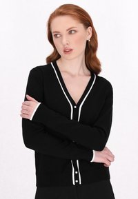 Cardigan negru cu margini albe, de tip V, cu mâneci lungi și detalii cu nasturi din perlă. Textură moale și design ajustat.