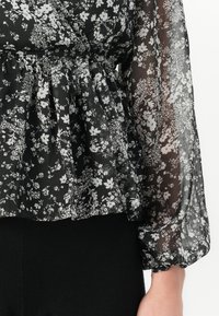 Blusa nera con stampa floreale bianca, tessuto trasparente, maniche lunghe a sbuffo e orlo con volant. Il materiale appare leggero e dalla texture interessante.