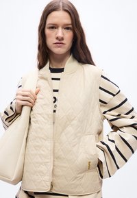 Νεαρή γυναίκα που φοράει ένα μπεζ γιλέκο με καπιτονέ λεπτομέρειες πάνω από μια μαύρη και λευκή ριγέ μπλούζα με μακριά μανίκια, κρατώντας μία τσάντα ώμου σε μπεζ χρώμα που ταιριάζει.