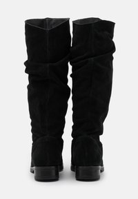 Une paire de bottes noires en suède montantes avec des talons blocs bas, montrées de dos sur un fond blanc.