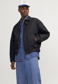 Giacca bomber nera con zip frontale, polsini elasticizzati e una tasca laterale. Indossata sopra una camicia di denim blu, abbinata a pantaloni di denim larghi.
