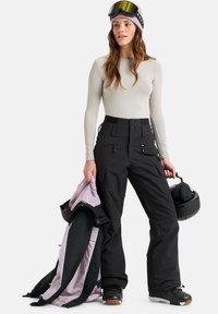 Black snow pants with a high waist and multiple pockets, paired with a long-sleeve light beige top. A purple and black jacket and helmet are held.