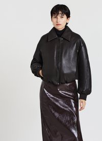 Veste bomber en cuir noir avec un large col, des poignets côtelés et une fermeture éclair, associée à une jupe longue et brillante de couleur marron foncé.