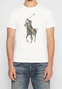 Bílé bavlněné tričko s barevným grafickým potiskem koně a jezdce, se šikmými liniemi v zelené, červené a žluté barvě. Volný střih.