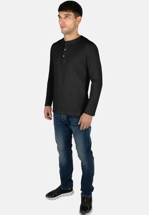 Black long-sleeve henley shirt with three-button placket, paired with blue jeans and black sneakers. Smooth fabric, fitted design.