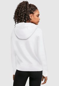 Felpa bianca con cappuccio, dal texture morbido, vista posteriore che mette in mostra un grande cappuccio, maniche lunghe e orlo dritto. Indossata con leggings neri.