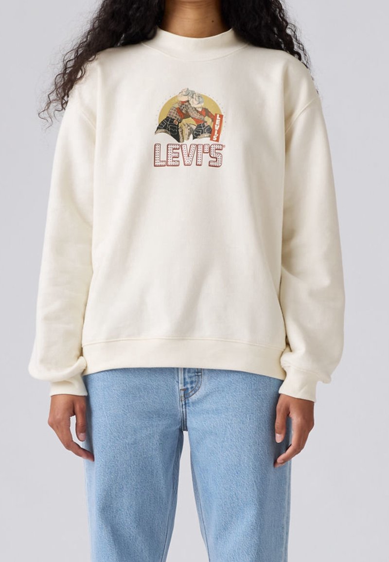 Personne portant un sweat-shirt Levi's écru avec un motif style vintage de deux cow-boys et un texte Levi's rouge, assorti à un jean bleu clair.