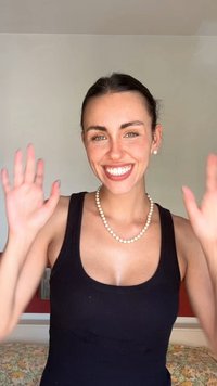 Black ribbed tank top, pearl necklace, smiling with hands raised. Light skin, freckles, and long dark hair pulled back. Neutral background.