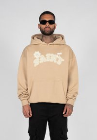 Sweatshirt beige à capuche avec le texte "SAINT" brodé en blanc et un design floral. Il dispose d'une poche avant et d'une coupe décontractée. Porté avec un pantalon cargo noir.