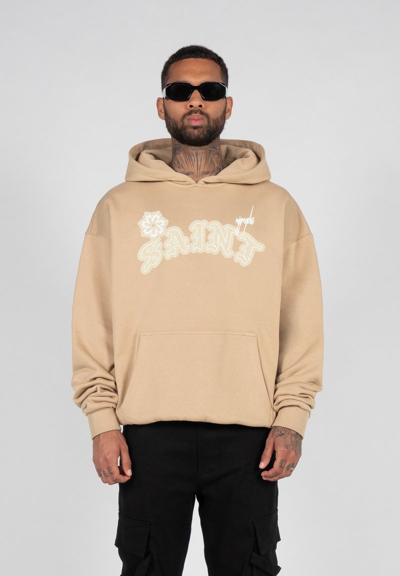 Sweatshirt beige à capuche avec le texte "SAINT" brodé en blanc et un design floral. Il dispose d'une poche avant et d'une coupe décontractée. Porté avec un pantalon cargo noir.