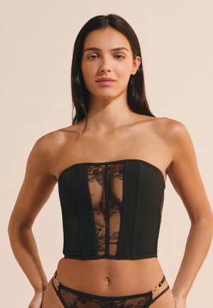 Donna con capelli lunghi e scuri indossa un top corsetto nero senza spalline con pannello in pizzo floreale e biancheria intima in pizzo coordinata su sfondo semplice.