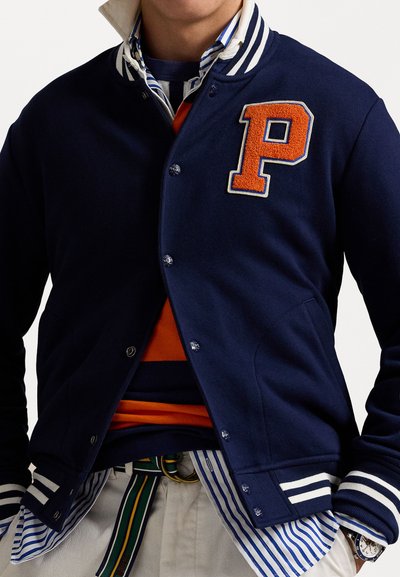 Námornícky vlnený varsity kabát s bielymi pruhovanými manžetami a golierom; na ňom sa nachádza oranžová plstenná nášivka "P". Nosený nad viacfarebnou pruhovanou košeľou.