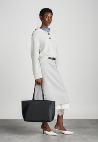 Μαύρη τσάντα tote με δομημένο σχέδιο, από λείο δέρμα, διπλά χερούλια και ανάγλυφο λογότυπο. Φοριέται με μια ανοιχτό γκρι φούστα και πουλόβερ.