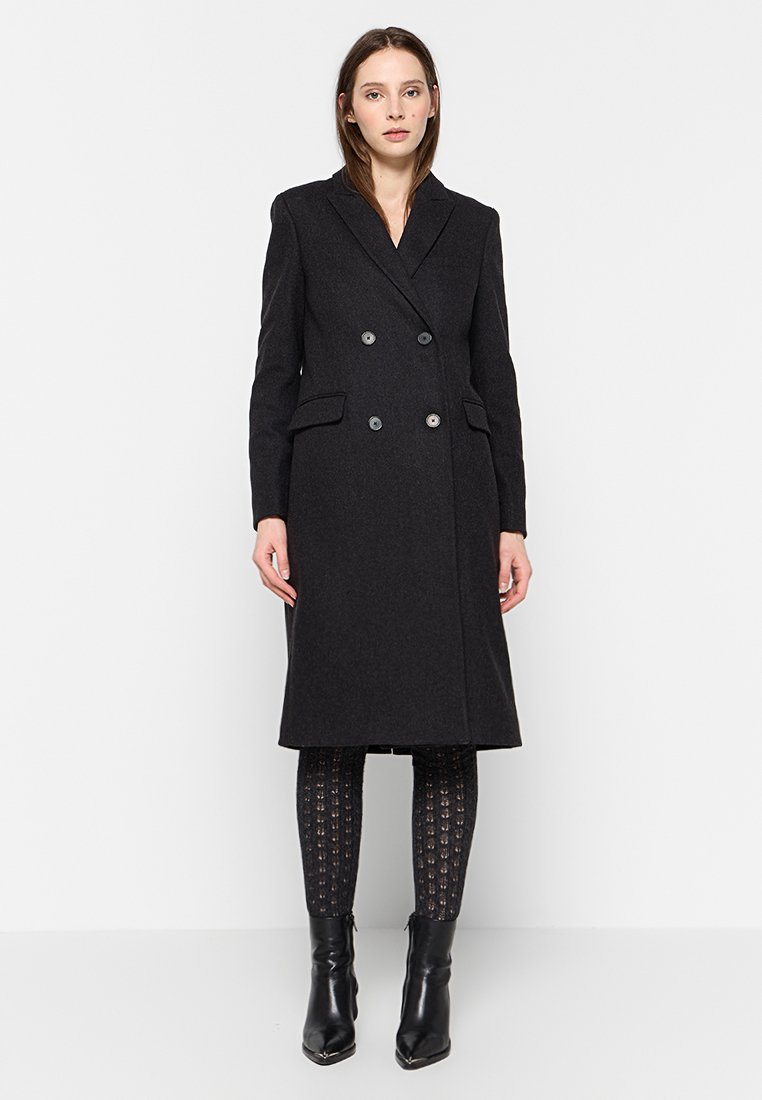 Manteau noir croisé avec un col cranté, des poches latérales et un tissu texturé, porté sur des leggings à motifs et des bottines.