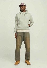 Homem a usar um hoodie cinza claro, boné bege e castanho, calças largas verde-oliva e botas bege, de pé com uma mão no bolso.