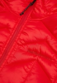 Rote Daunenjacke mit gestepptem Design, ausgestattet mit einem Reißverschluss und dem in dunklerem Rot gestickten "PeakPerformance"-Logo. Glatte, glänzende Textur.