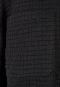 Tissu noir texturé avec un motif en grille montrant une couture horizontale et un détail de poche arrondi sur un vêtement.