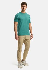 T-shirt teal a collo qui presenta una tasca sul petto, abbinato a pantaloni tapered beige chiaro e sneakers grigie. Look casual con una vestibilità rilassata.