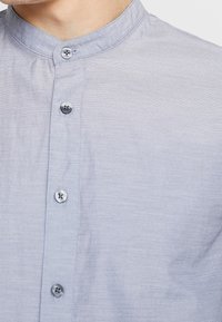 Ljusblå skjorta med knäppning och ståkrage, textil med struktur och runda silverknappar längs framsidan. En enkel och stilren design.