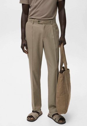 Man met beige broek, bijpassende sandalen en een korte mouwen overhemd, houdt een bruine draagtas naast zich vast.