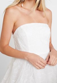 Robe blanche sans bretelles avec un tissu texturé, présentant des motifs en couches et en vagues ainsi que des sequins. Design élégant avec un corsage ajusté et une jupe évasée.