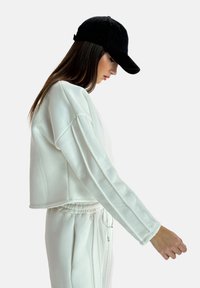 Witte cropped sweatshirt met lange mouwen van een textuurrijke, zachte stof. Gekenmerkt door een trekkoord en witte broek, gecombineerd met een zwarte baseballpet.