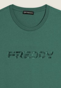 Camiseta de algodón verde con cuello redondo. Presenta un diseño de texto "FREDDY" en estilo recortado que revela un patrón de fondo texturizado.