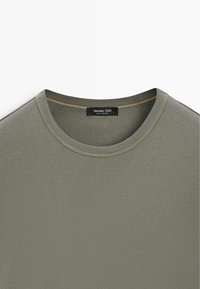 T-shirt vert olive à col rond avec détail de couture beige et une étiquette noire indiquant "Massimo Dutti Made in Portugal" au niveau de l'encolure.