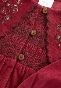 Robe en velours côtelé rouge avec des motifs floraux brodés, un corsage smocké, des boutons bruns et des détails de décolleté festonné.