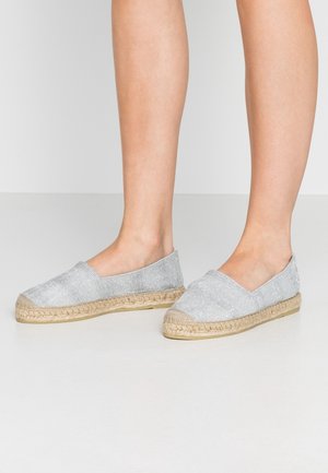 Sølvfarvede slip-on espadrille-sko med flettede jutesåler, båret på bare fødder mod en ensartet lys baggrund.