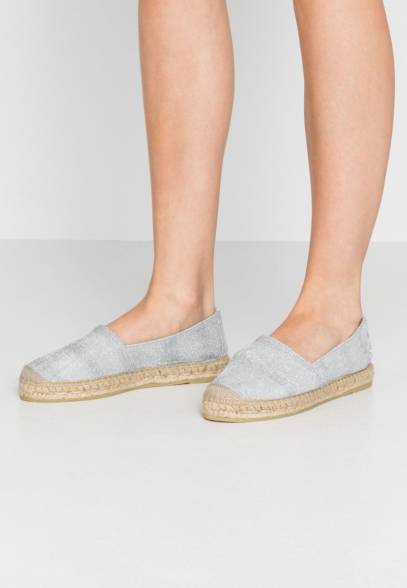 Espadrilles argentées à enfiler avec semelles en jute tissé portées pieds nus sur un fond clair uni.