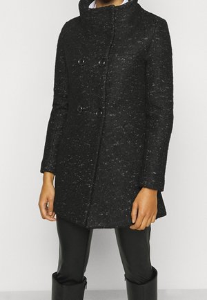 Persona que lleva un abrigo negro de doble botonadura con textura y botones grandes, combinado con pantalones negros y botas negras hasta la rodilla.