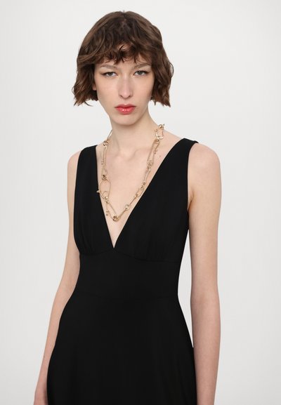 Zwarte jurk met een diepe V-halslijn, getailleerde lijfje en vloeiende rok. Gecombineerd met een gouden ketting met ovale schakel.