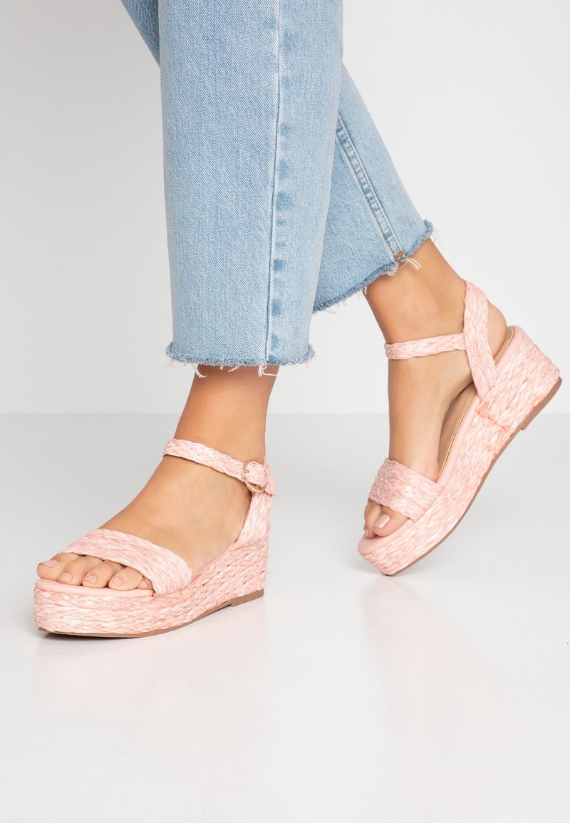 Sandales à talons compensés en rose doux, avec une texture tissée, de larges brides et une boucle à la cheville. Portées avec un jean court bleu clair.