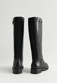 Botas negras hasta la rodilla que presentan un frente de cuero suave y paneles laterales elásticos. Incluyen un tacón bajo y apilado con un detalle metálico.
