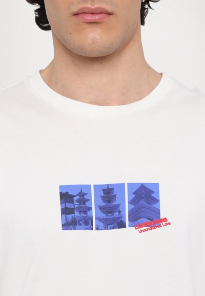 T-shirt en coton blanc avec un graphique bleu rectangulaire représentant trois structures architecturales et le texte "LES BEAUMANS Amour Inconditionnel" en rouge.