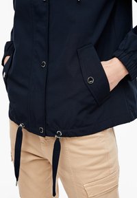 Chaqueta azul marino con herrajes plateados, cordones en el dobladillo y puños elásticos. Pantalones cargo beige con bolsillos laterales. Tela suave y texturizada.